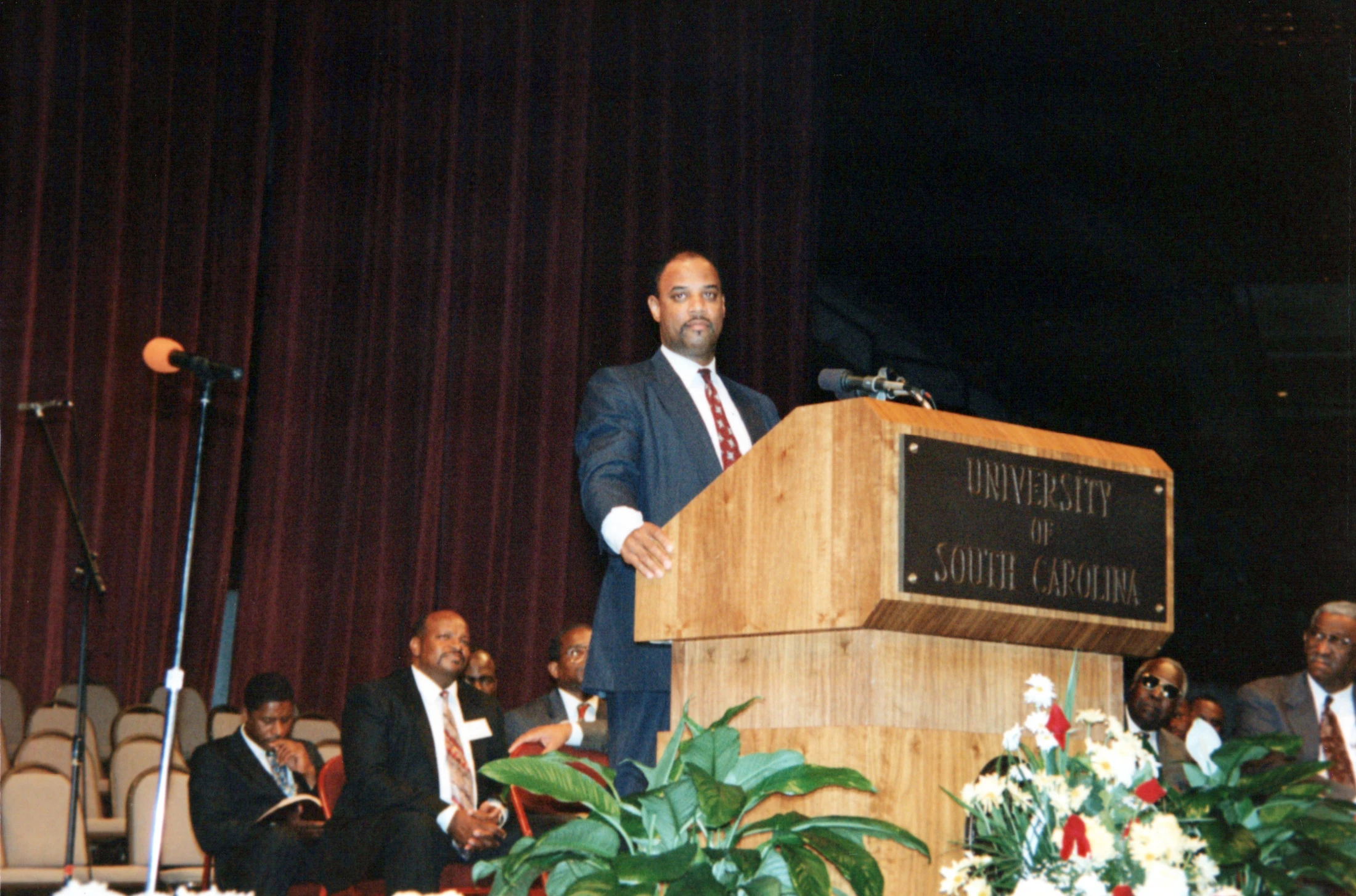 A man in a suit stands at a wooden podium with a 'University of South Carolina' plaque, addressing an audience. Other men in suits are seated nearby.