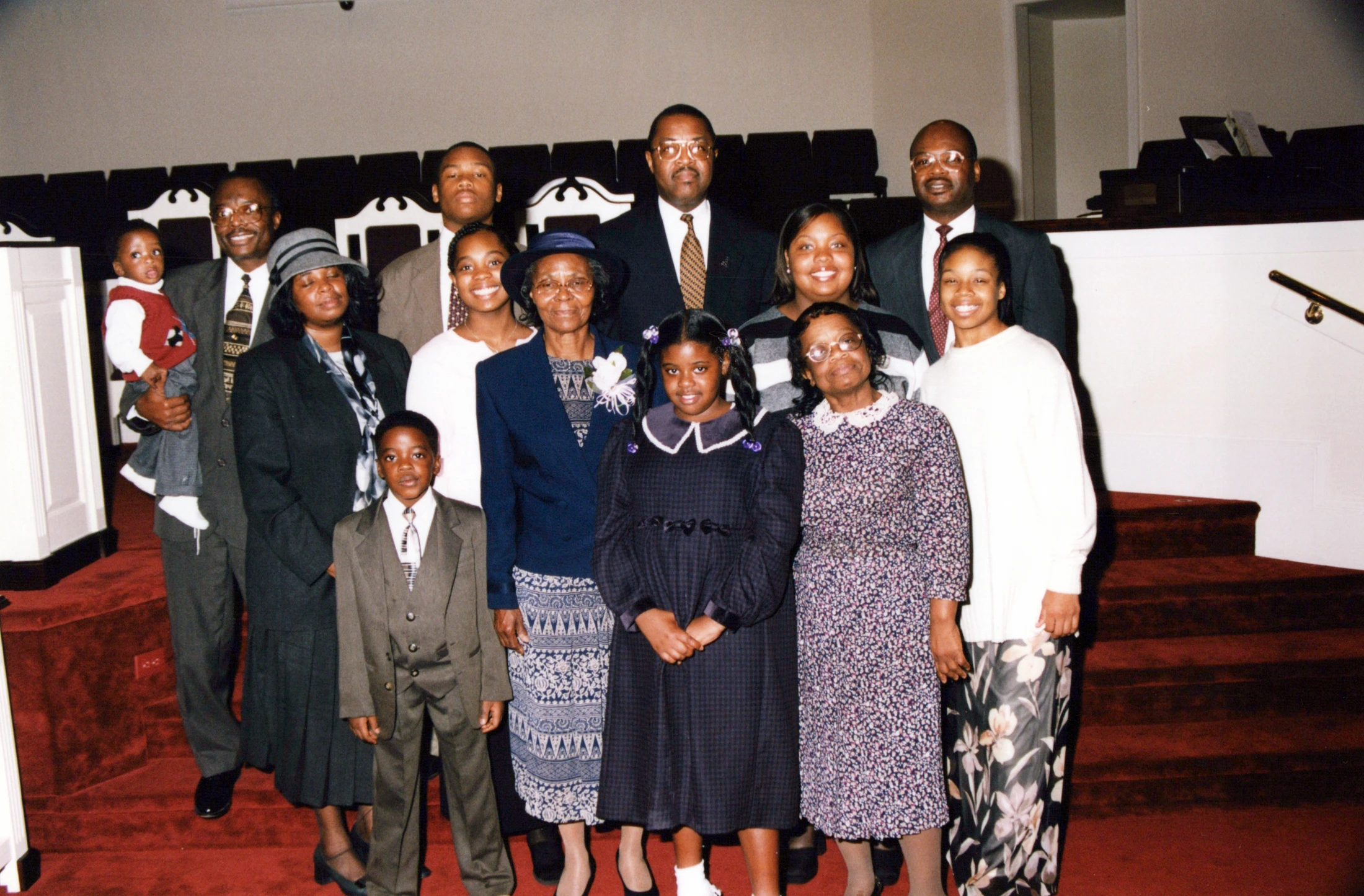 A large African American family with multiple generations, dressed in formal attire, poses for a group photo on the red carpeted steps inside a church.