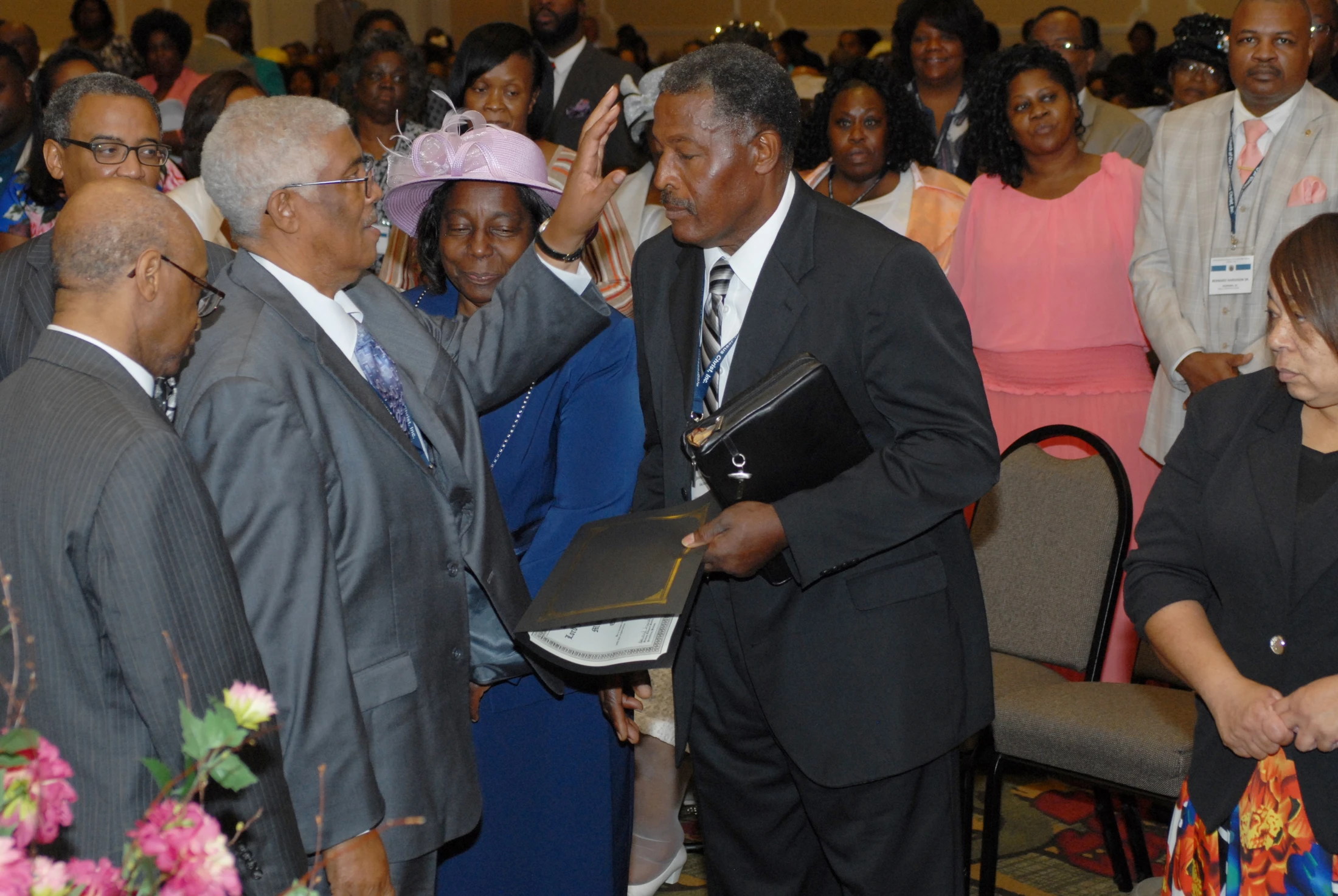 An elderly Black man in a suit blesses another man during a formal gathering, surrounded by other attendees watching the interaction.