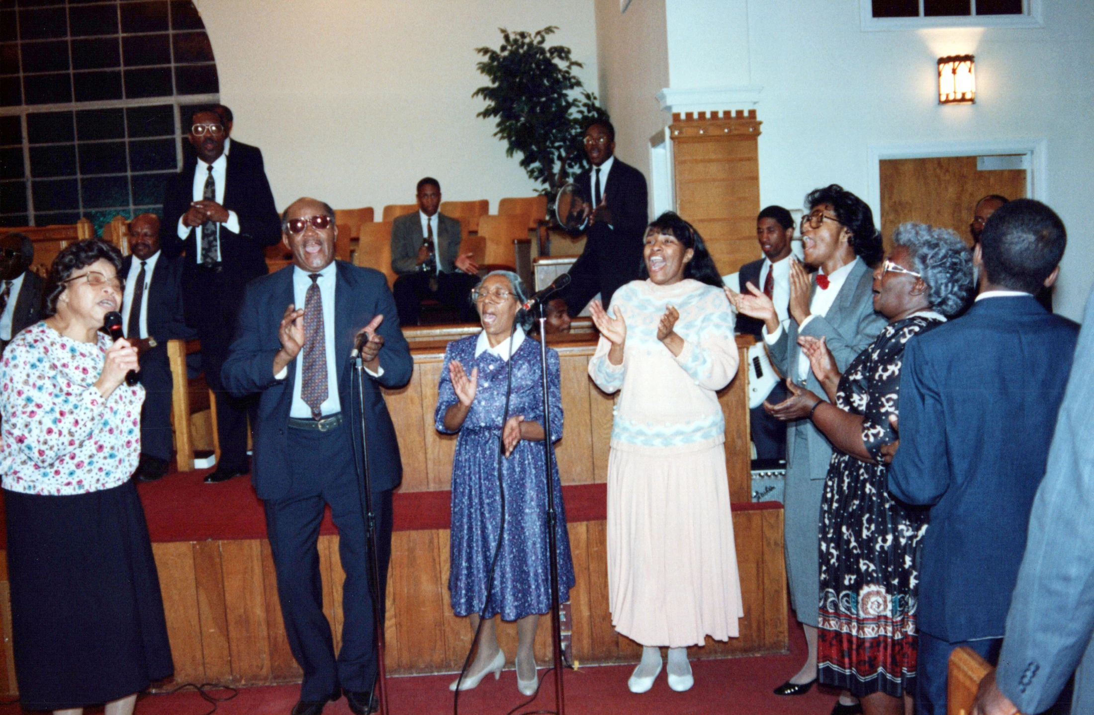 A vintage photograph of a Black church choir with men and women singing and clapping with joy on a wooden stage with a red carpet.