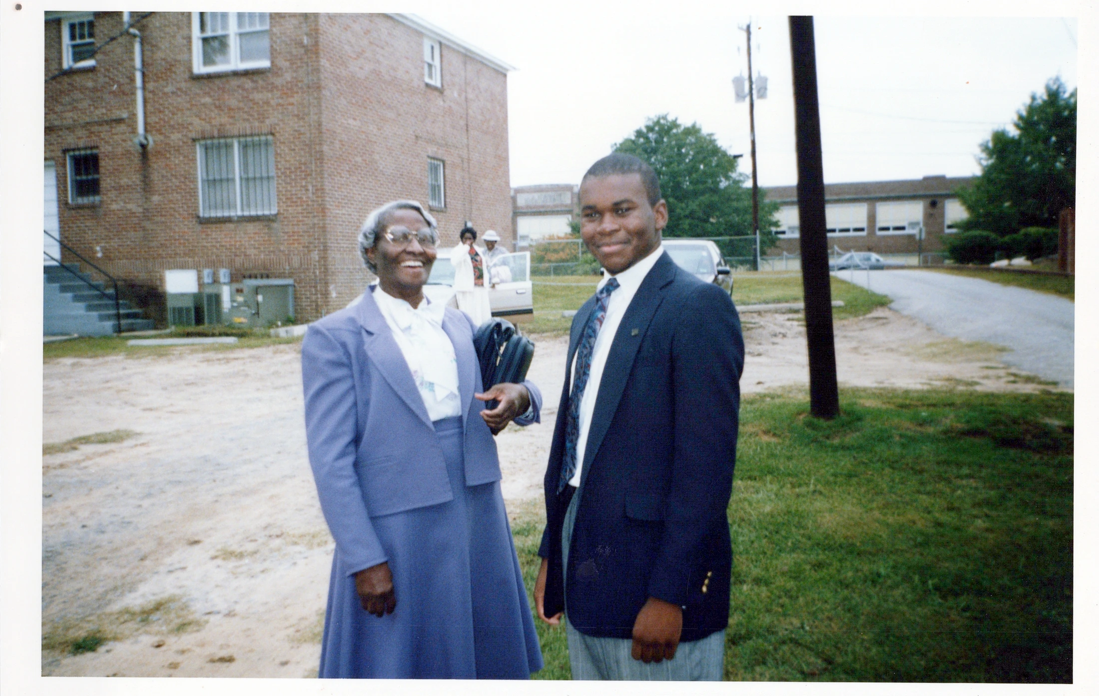 An outdoor portrait of an older Black woman in a blue suit and a younger Black man in a blazer, both smiling at the camera in front of a brick building.