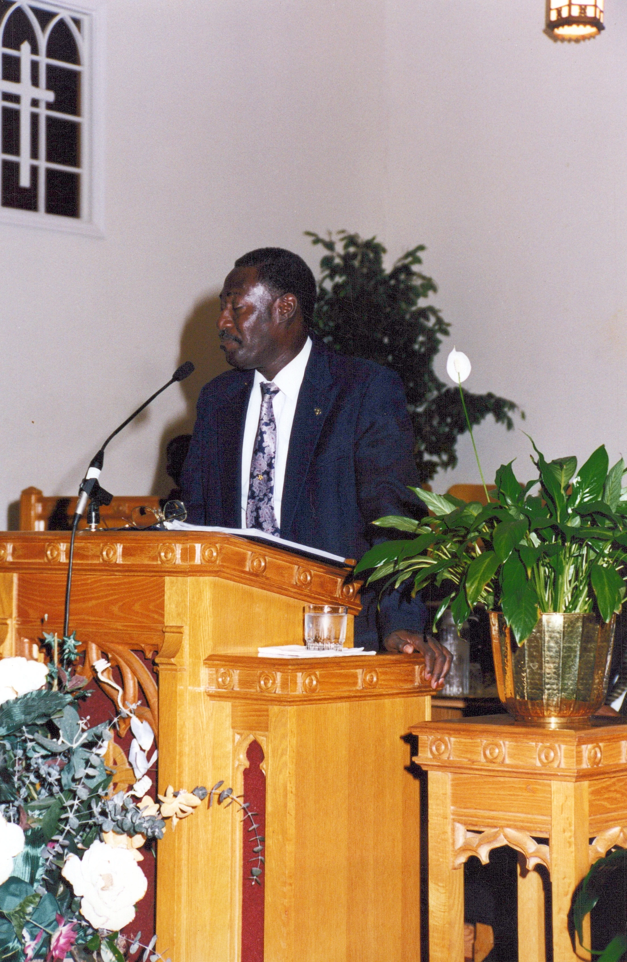 A Black man in a dark suit and patterned tie stands behind a wooden pulpit in a church, speaking into a microphone with his eyes closed or looking down.