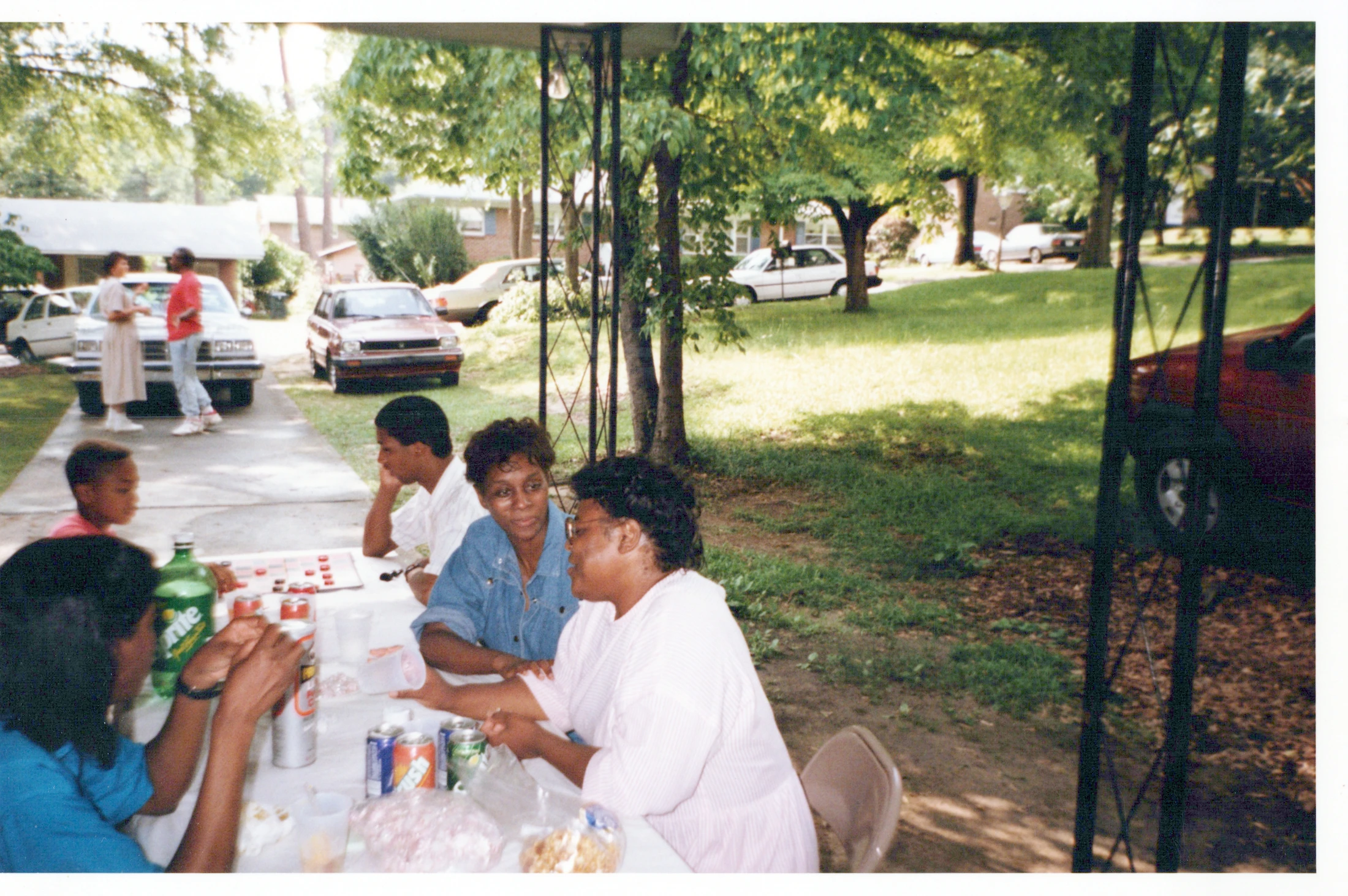 A vintage photograph of a Black family enjoying an outdoor gathering. They sit at a table with drinks, talking, with trees and cars in the background.