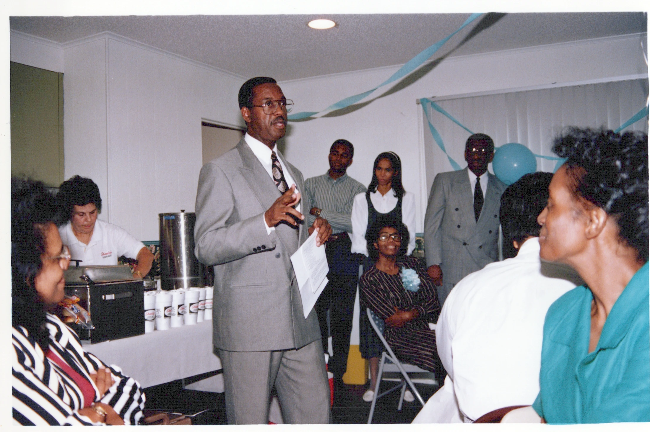 An African American man in a grey suit and glasses speaks to a group of people at an indoor event decorated with blue streamers and balloons.