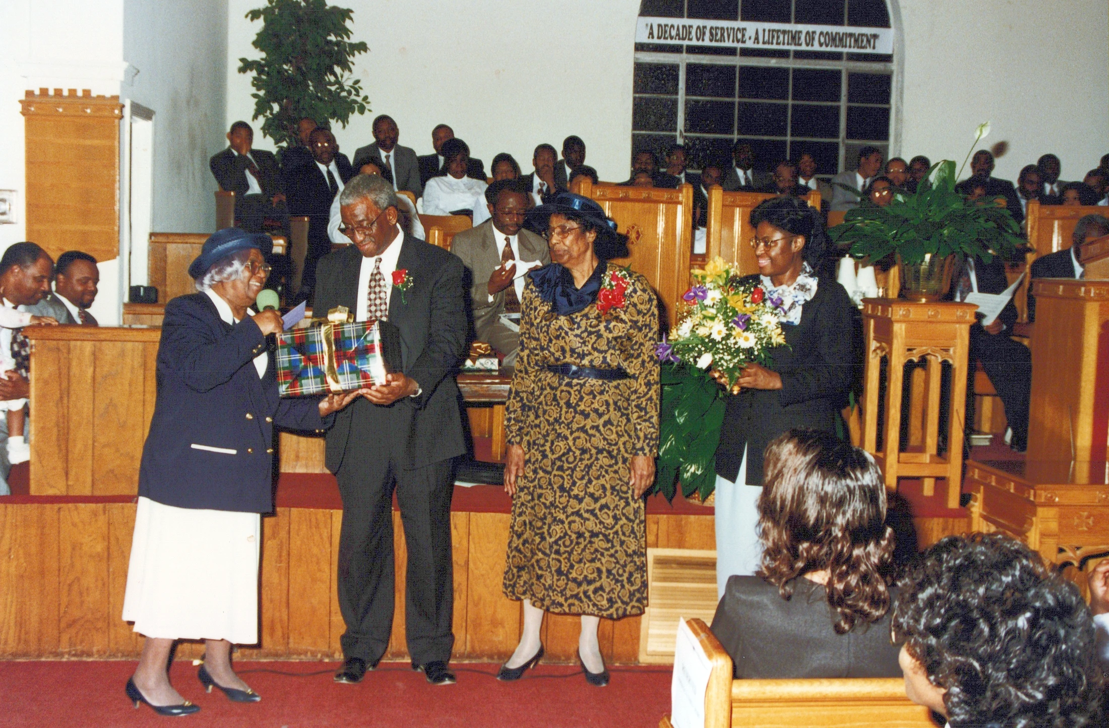 At the front of a church, an older Black woman presents a wrapped gift to a man in a suit, while another woman holds flowers. The congregation looks on.