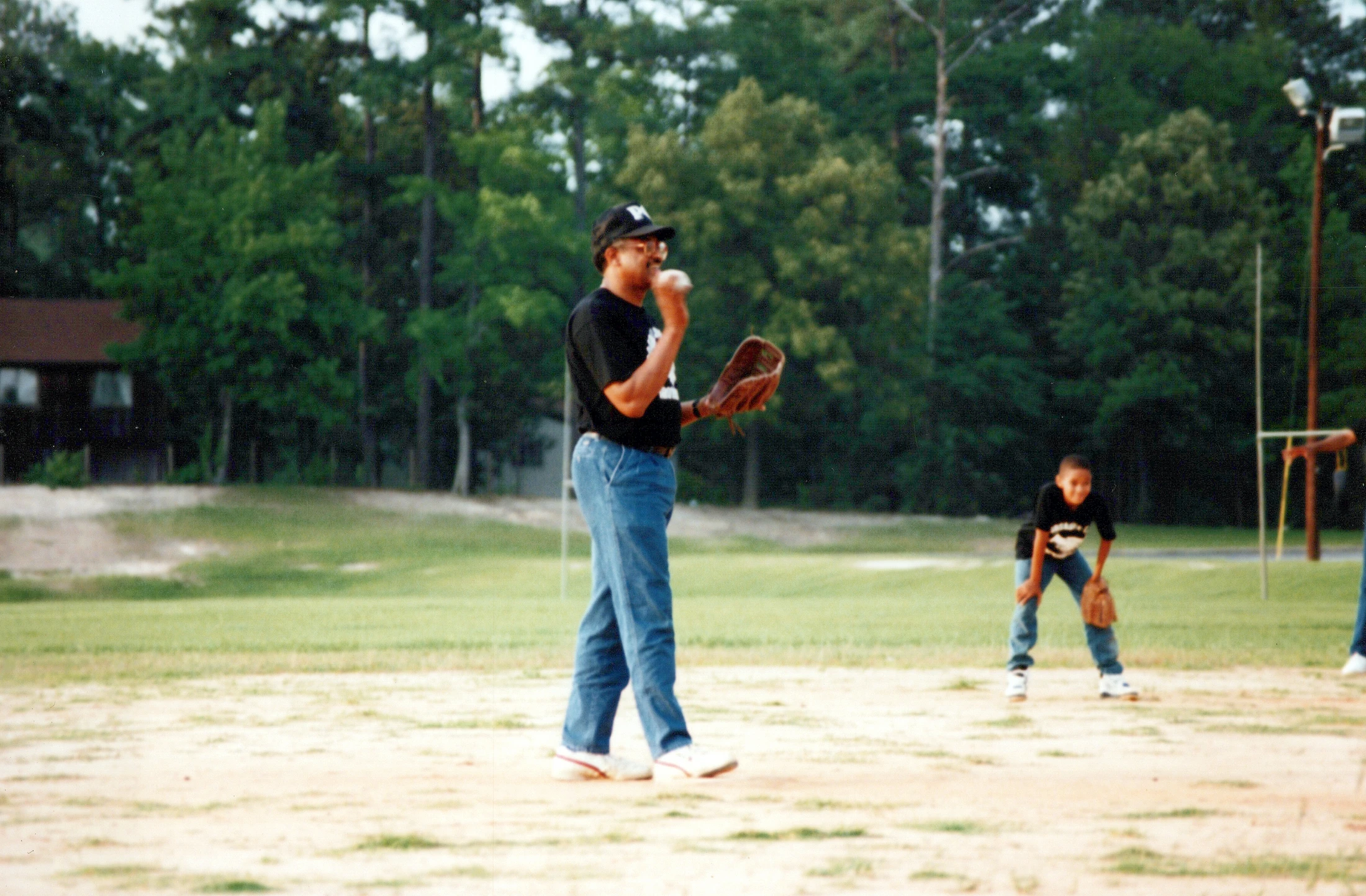 A vintage photo of a man in a black shirt and a baseball cap about to throw a baseball to a young boy on a dirt and grass field.