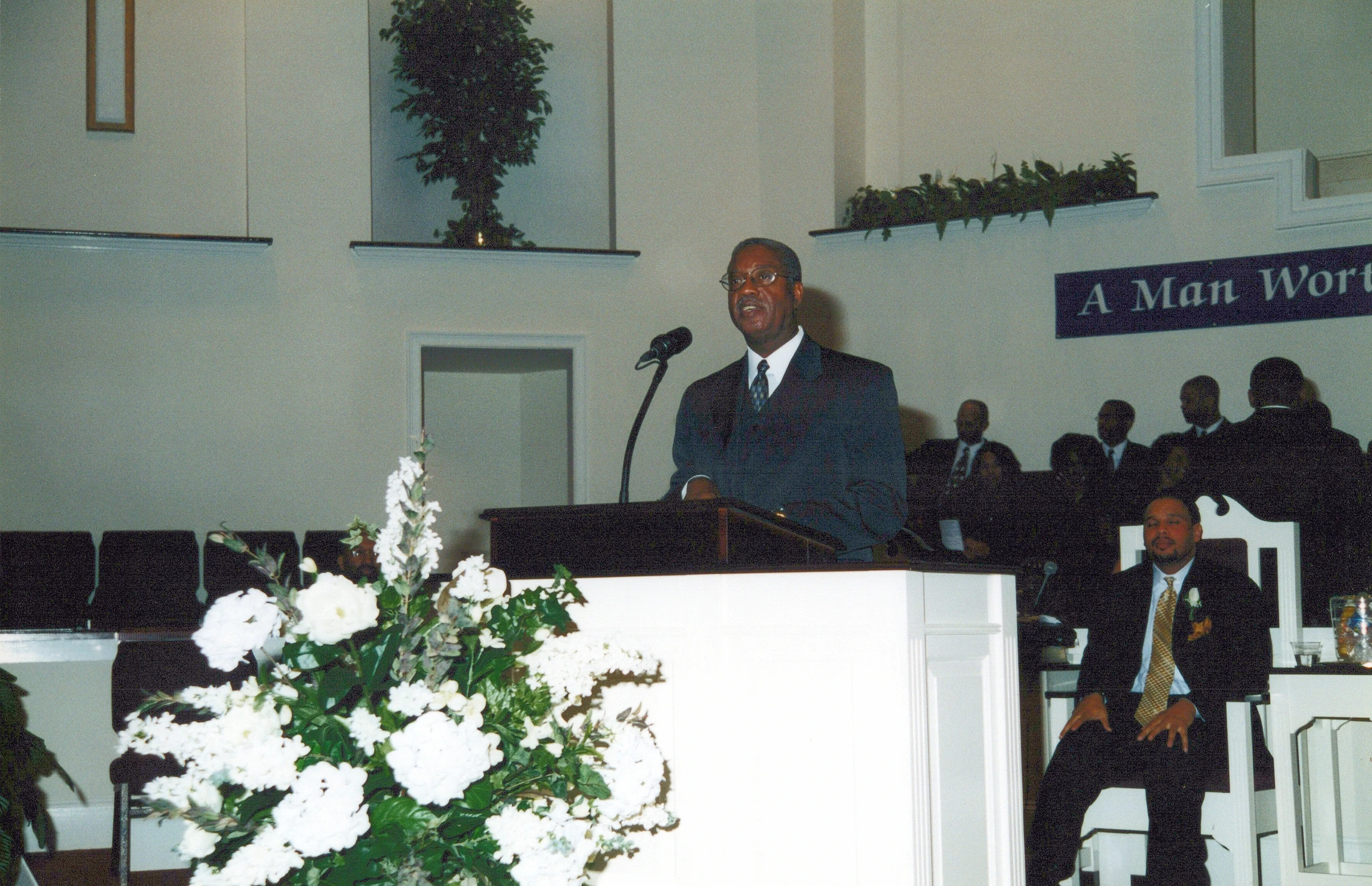 A Black man in a dark suit and glasses speaks at a white podium in a church, with a large white floral arrangement in the foreground and an audience behind him.