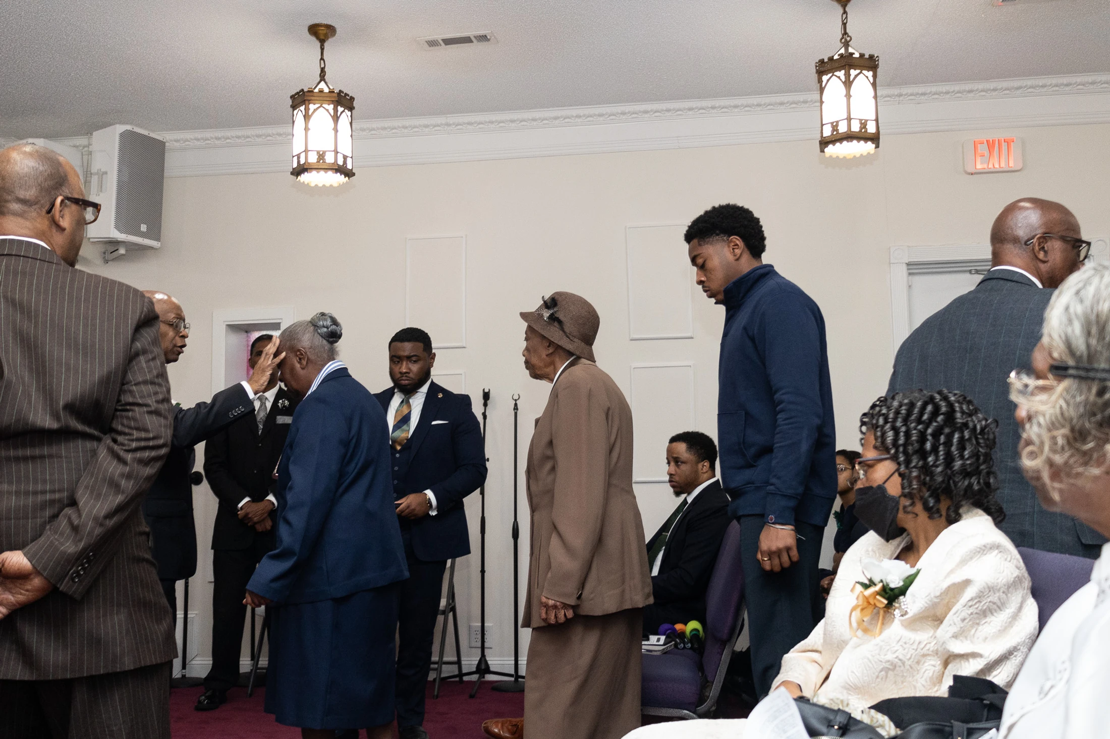 Inside a church, a group of formally dressed Black people are gathered. A man blesses an elderly woman by placing his hand on her forehead as others watch on.