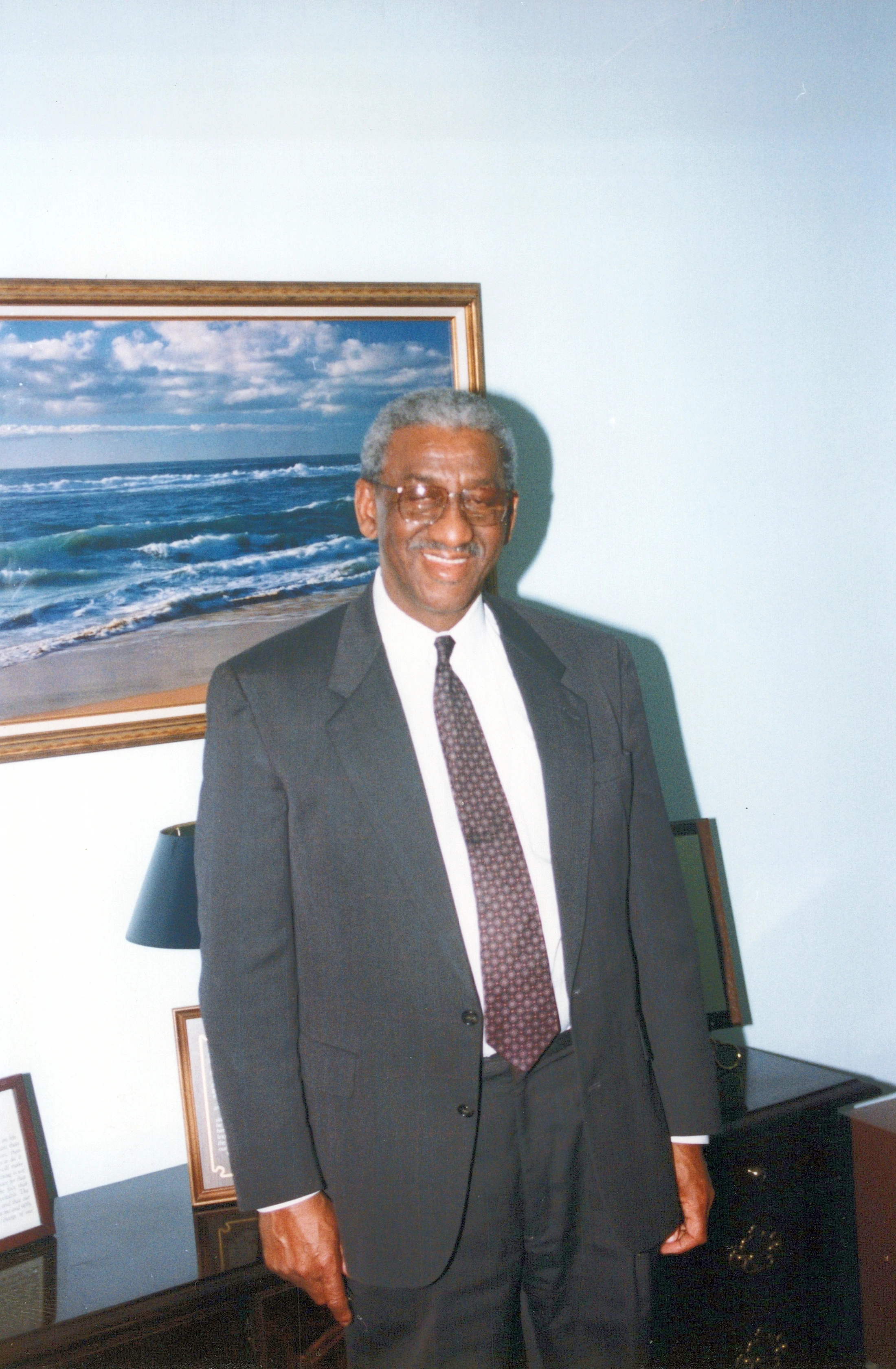 An older African American man with gray hair and glasses smiles while wearing a dark suit and tie. He stands in an office in front of a painting of the ocean.