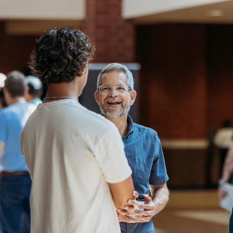 Three men of different ages stand together in a lobby, engaged in conversation. The man in the center wears glasses and smiles warmly at the others.