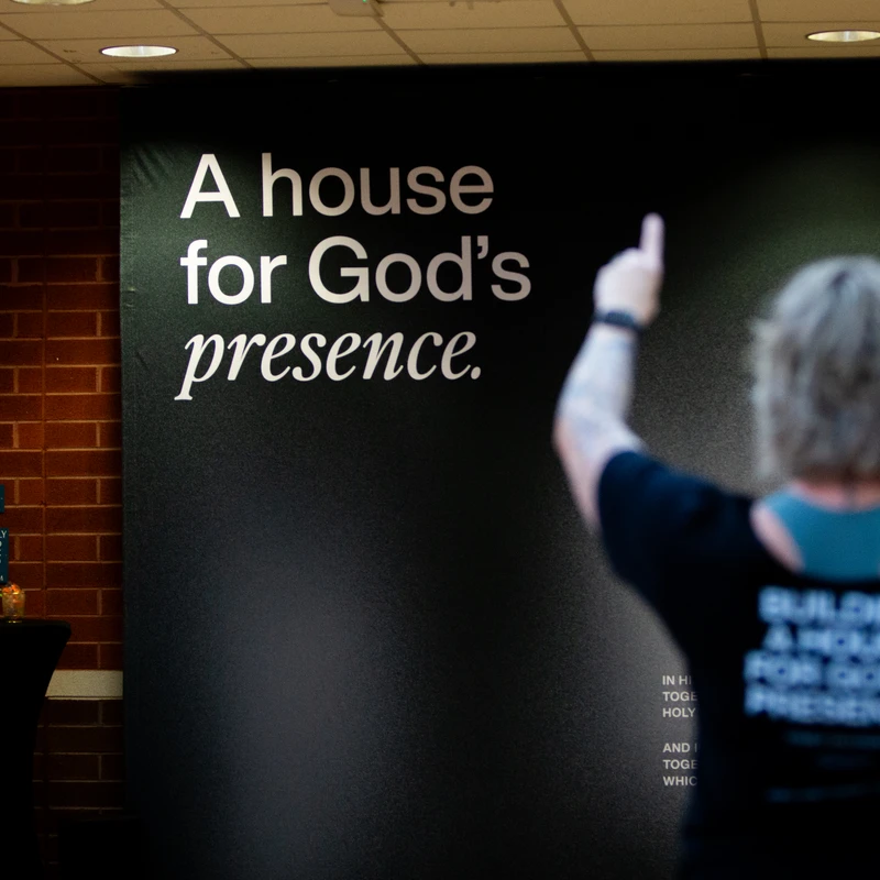 A person, viewed from behind, points up at a large black banner with the white text 'A house for God's presence' against a brick wall.