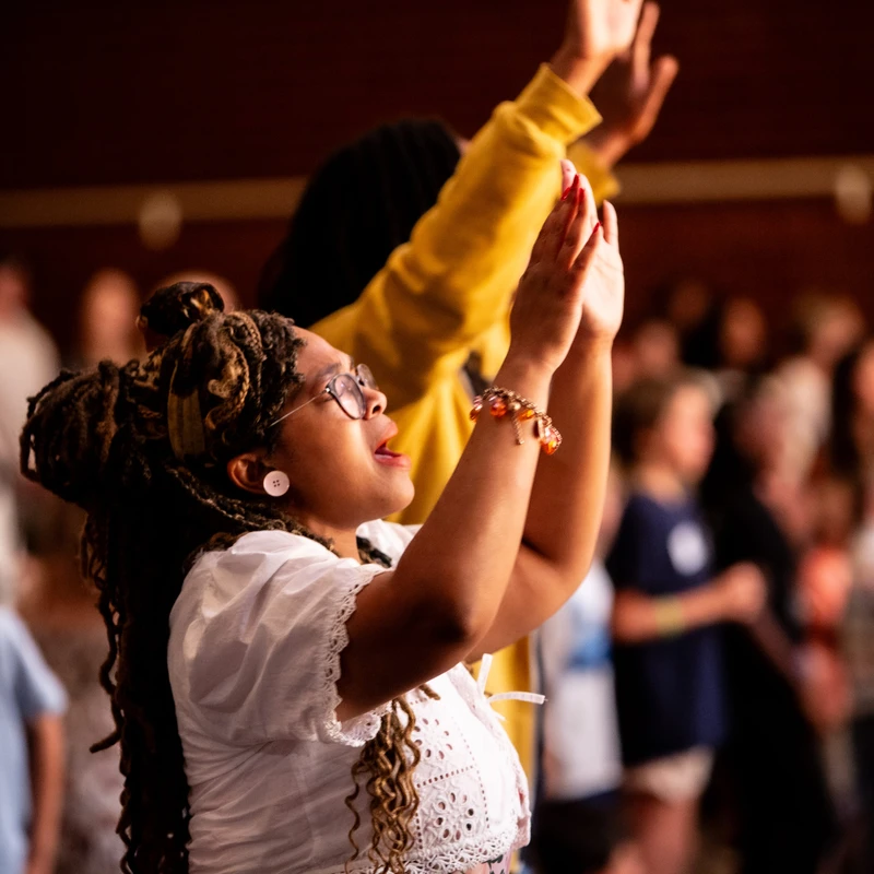 A Black woman with dreadlocks and glasses, wearing a white top, stands in a crowd with her hands raised in worship, looking up with a devoted expression.