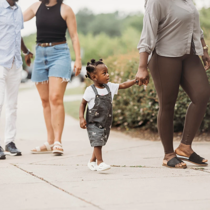 A young Black girl in denim overalls holds an adult's hand as she walks on a sidewalk with a group of people outdoors.