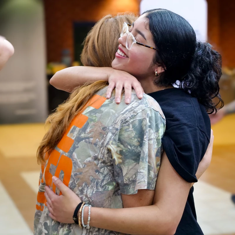 A young woman with dark hair and glasses smiles warmly as she hugs a friend with reddish-brown hair wearing a camouflage t-shirt.