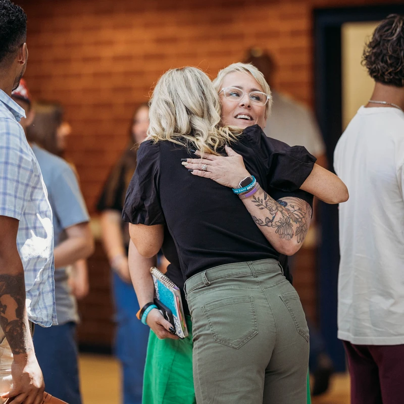 A blonde woman with glasses and a floral tattoo smiles as she warmly hugs another woman in a crowd of people during a social gathering.