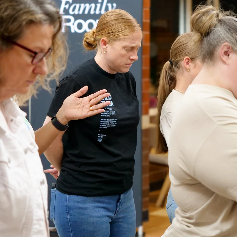 A woman with her eyes closed in prayer stands while another woman to her left raises a hand towards her in a gesture of blessing during an indoor event.
