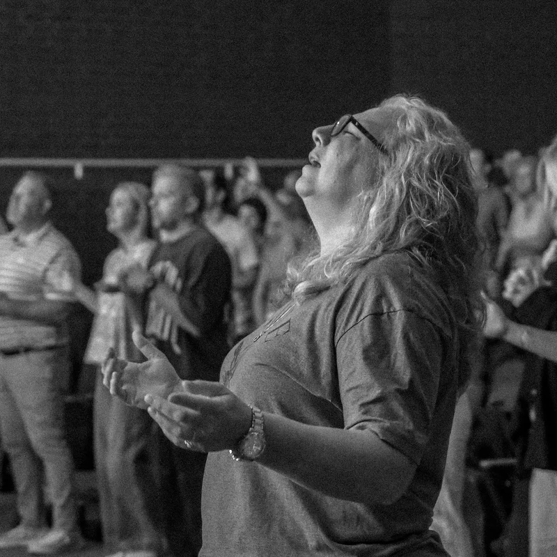 A black and white photo of a woman looking up with her hands open in prayer, surrounded by other people during a church worship service.