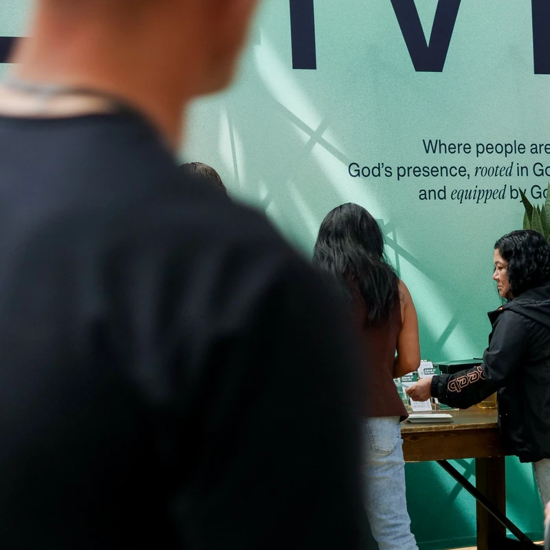 From behind a blurred man, two women stand at a welcome table in a church lobby, in front of a large green banner with religious text.
