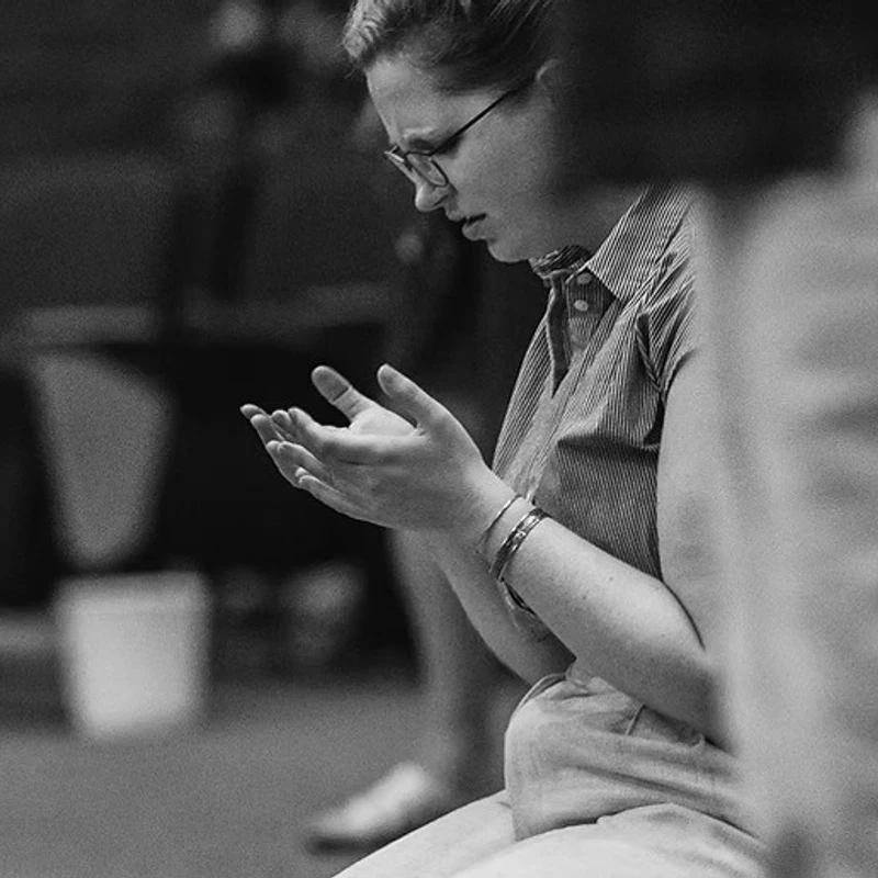 A black and white side-view photo of a woman with glasses praying. Her hands are open and her expression is solemn as she looks down.