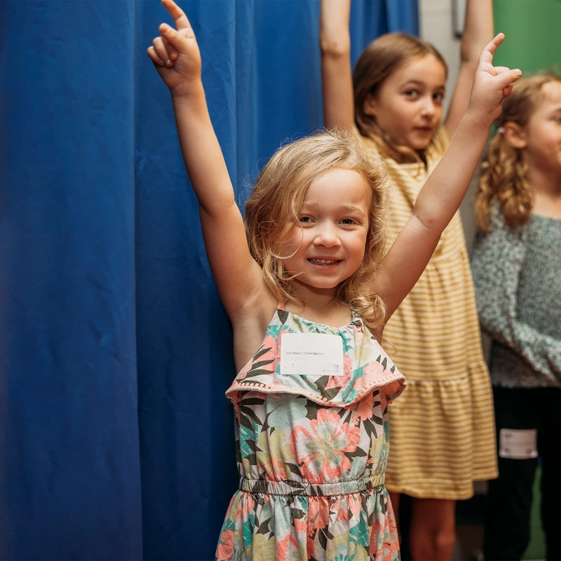 A young blonde girl in a floral sundress smiles at the camera with her arms raised in the air, standing with other children in front of a blue curtain.