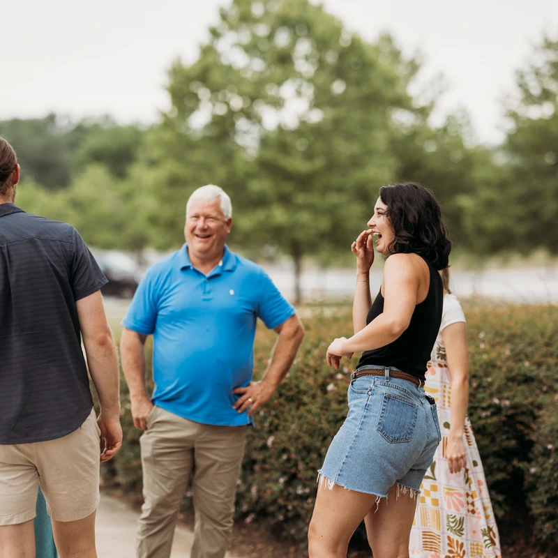 A group of people stand outdoors talking. A woman in a black tank top and jean shorts is laughing, while an older man in a blue polo shirt smiles.