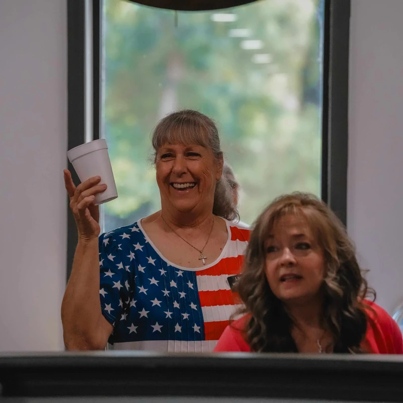 A smiling woman with graying hair, wearing an American flag shirt, holds up a white cup while standing under a lit sign of a coffee cup in a cafe.
