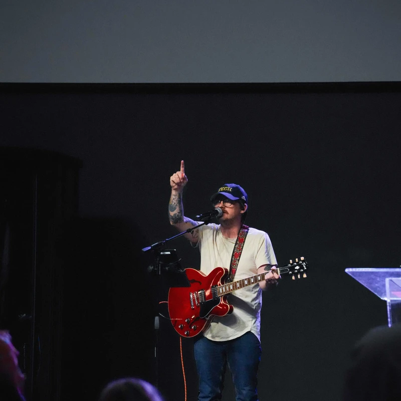 A man in a cap and glasses plays a red electric guitar and sings on a dark stage. He points a finger up, with lyrics on a screen behind him.