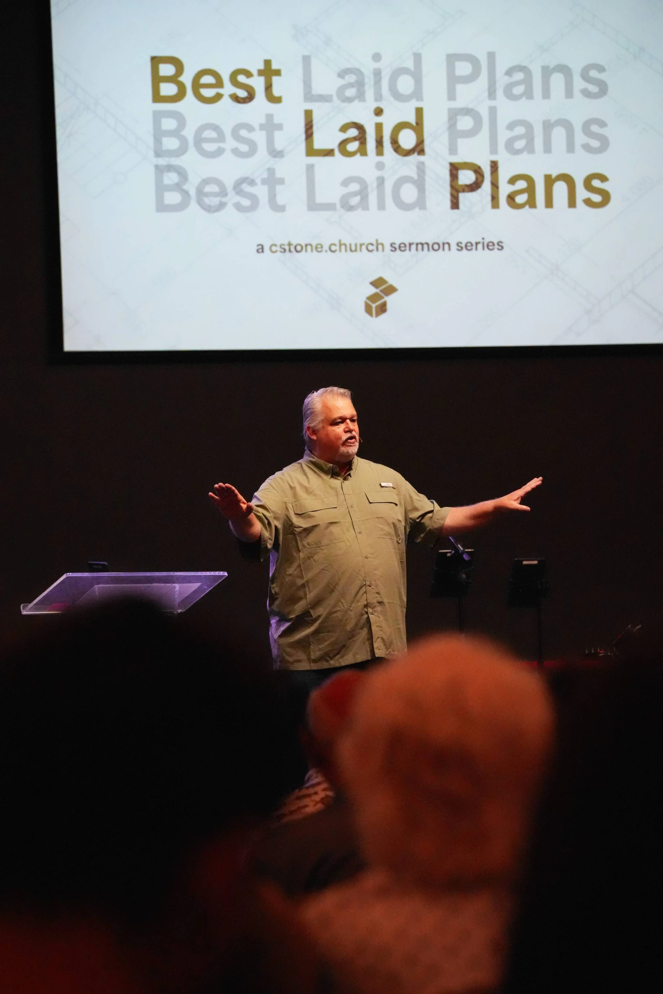 A gray-haired man in a green shirt stands on a stage, speaking. Behind him, a screen reads "Best Laid Plans, a cstone.church sermon series."