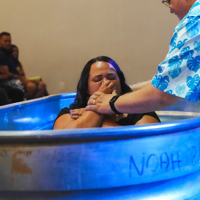 A pastor prepares to baptize a woman in a metal tub filled with blue-lit water. The woman has her eyes closed and a hand over her nose in anticipation.