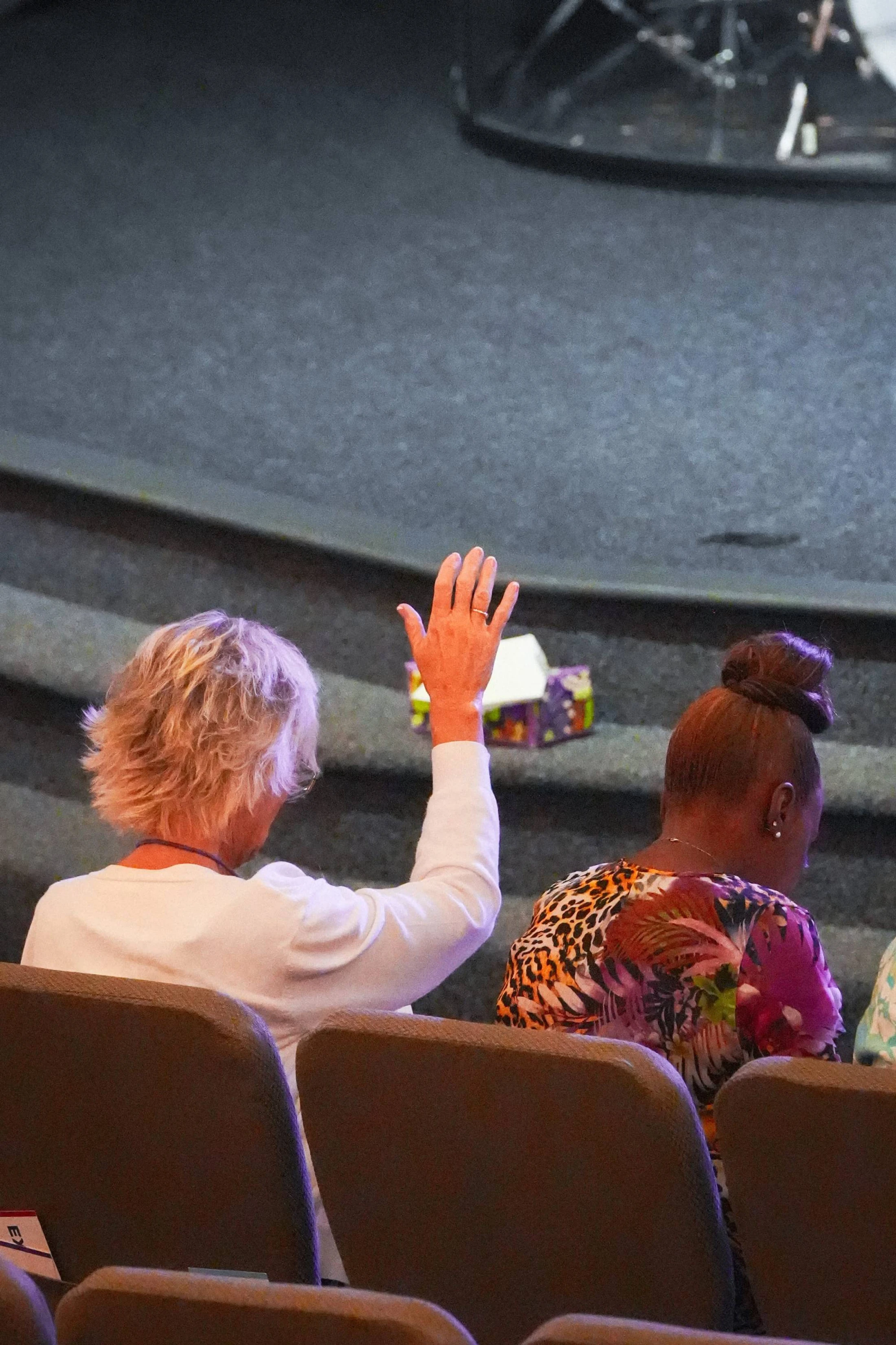From behind, a woman with short blonde hair in a white shirt raises her hand in worship while sitting in an auditorium next to another woman.