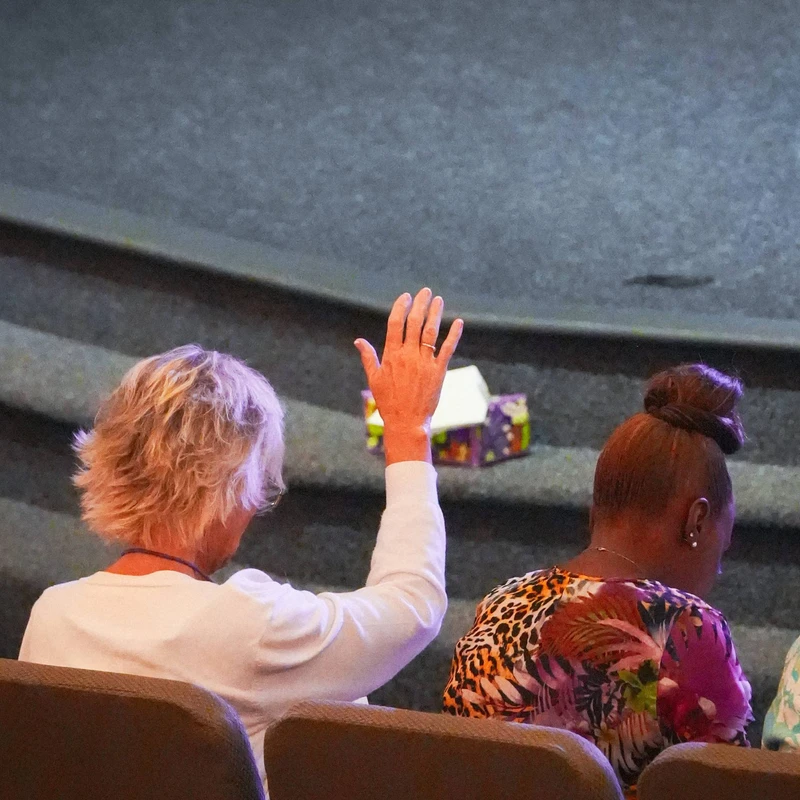 From behind, a woman with short blonde hair in a white shirt raises her hand in worship while sitting in an auditorium next to another woman.