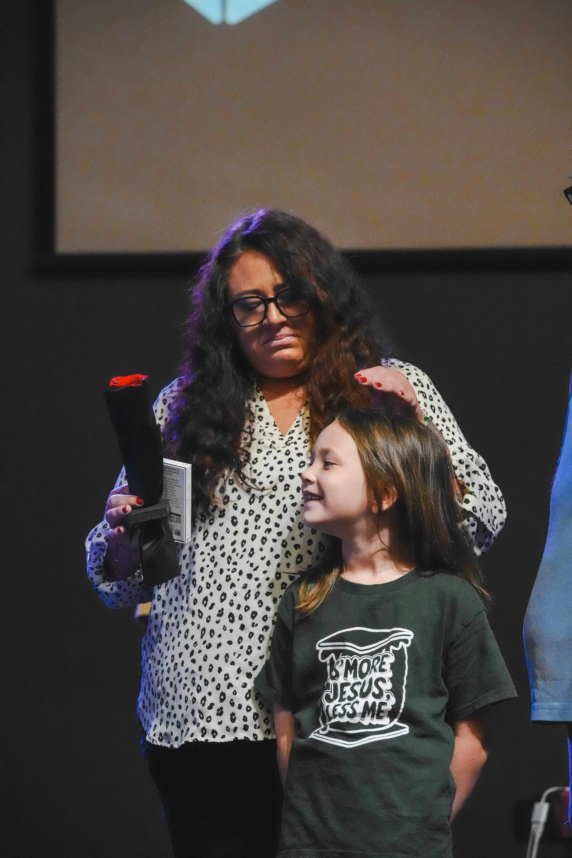 A woman in a spotted blouse stands behind a smiling young girl in a green t-shirt that says 'More Jesus Less Me,' gently touching her hair.