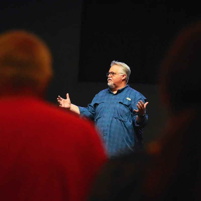 A man with gray hair and a beard speaks on stage, gesturing with his hands. The shot is from the audience's perspective with blurred figures in the foreground.