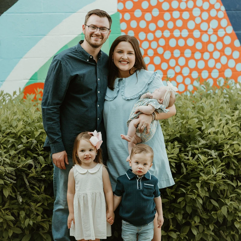 A smiling family of five, with a man, woman, two young children, and a baby, pose for a portrait in front of a colorful outdoor mural and green bushes.