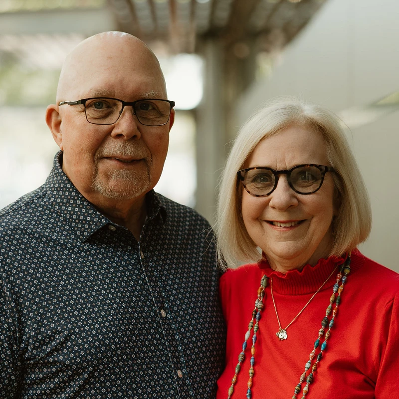 A friendly older couple smiles for the camera. The man is bald with a goatee and glasses, and the woman has blonde hair, glasses, and wears a red top.