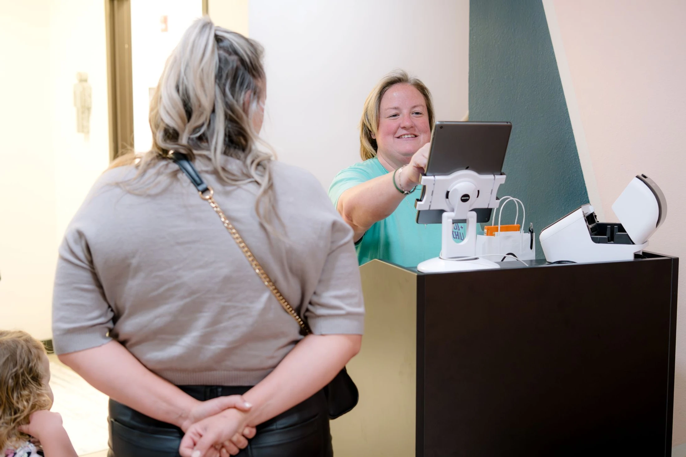 A smiling female receptionist in a mint green shirt helps a customer, pointing to a tablet screen at a modern front desk while the customer looks on.