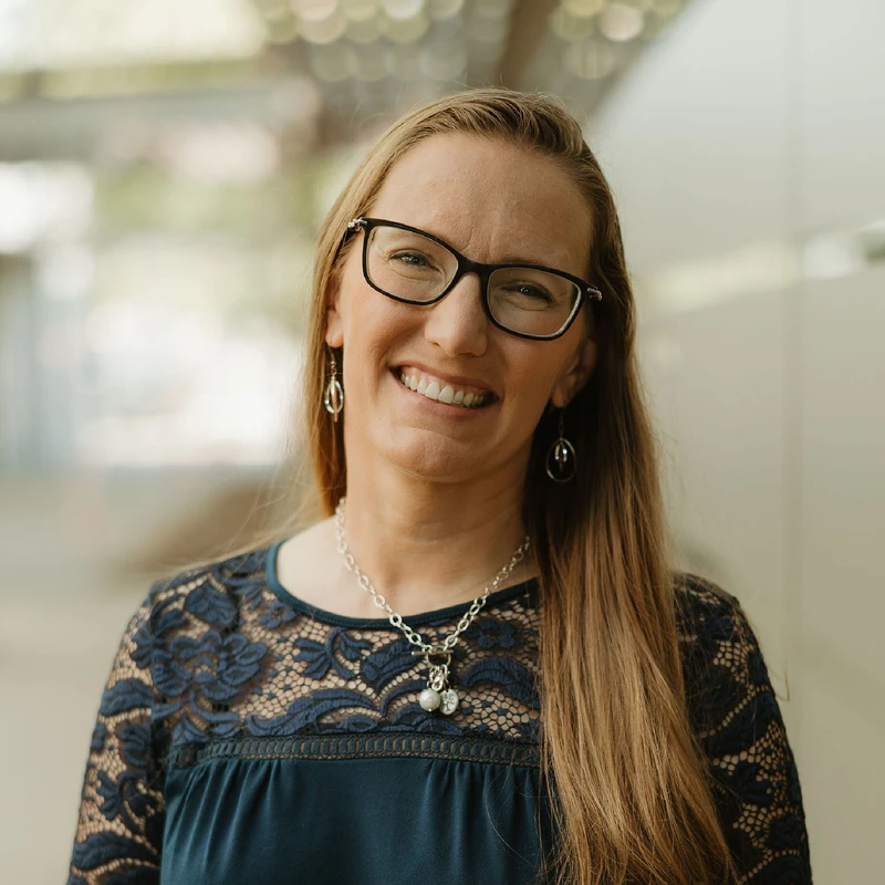A headshot of a smiling woman with long blonde hair and black-rimmed glasses, wearing a navy blue lace top and a silver charm necklace.