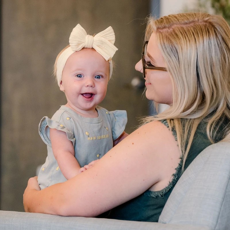 A blonde woman holds a smiling baby girl wearing a large cream bow. The baby looks at the camera while the woman gazes lovingly at her.