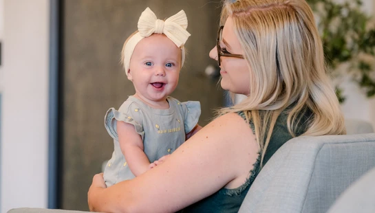 A blonde woman holds a smiling baby girl wearing a large cream bow. The baby looks at the camera while the woman gazes lovingly at her.