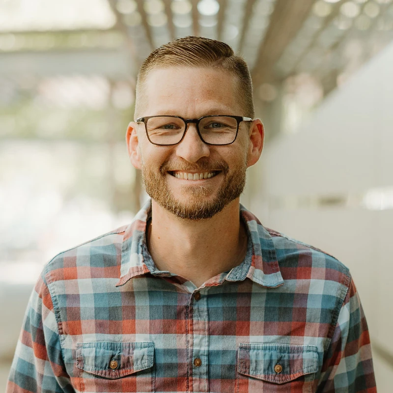 A friendly-looking man with short brown hair, a beard, and glasses smiles at the camera. He is wearing a red and blue plaid button-down shirt.
