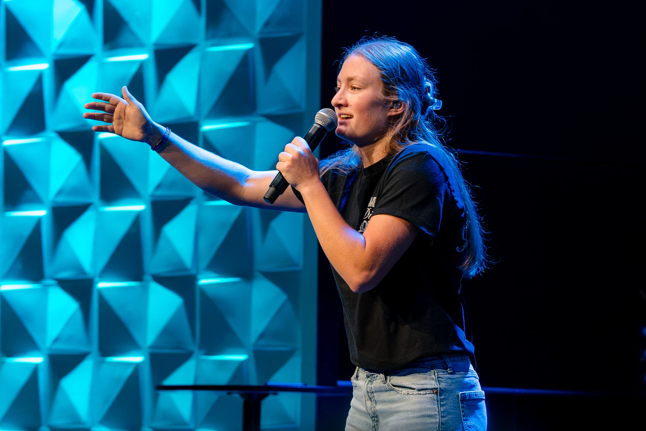 A young woman with long hair speaks into a microphone on a stage. She gestures with one hand against a backdrop of geometric blue panels and dark background.