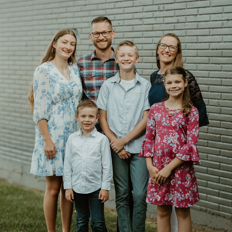A family of six, with parents and four children, smiles for a portrait while standing outside in front of a painted grey brick wall.