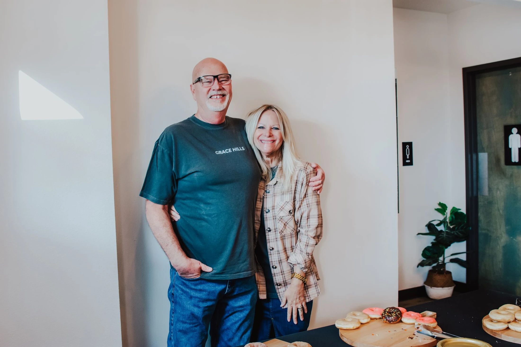 A smiling man and woman pose with their arms around each other, standing behind a table laid out with fresh donuts at an indoor gathering.