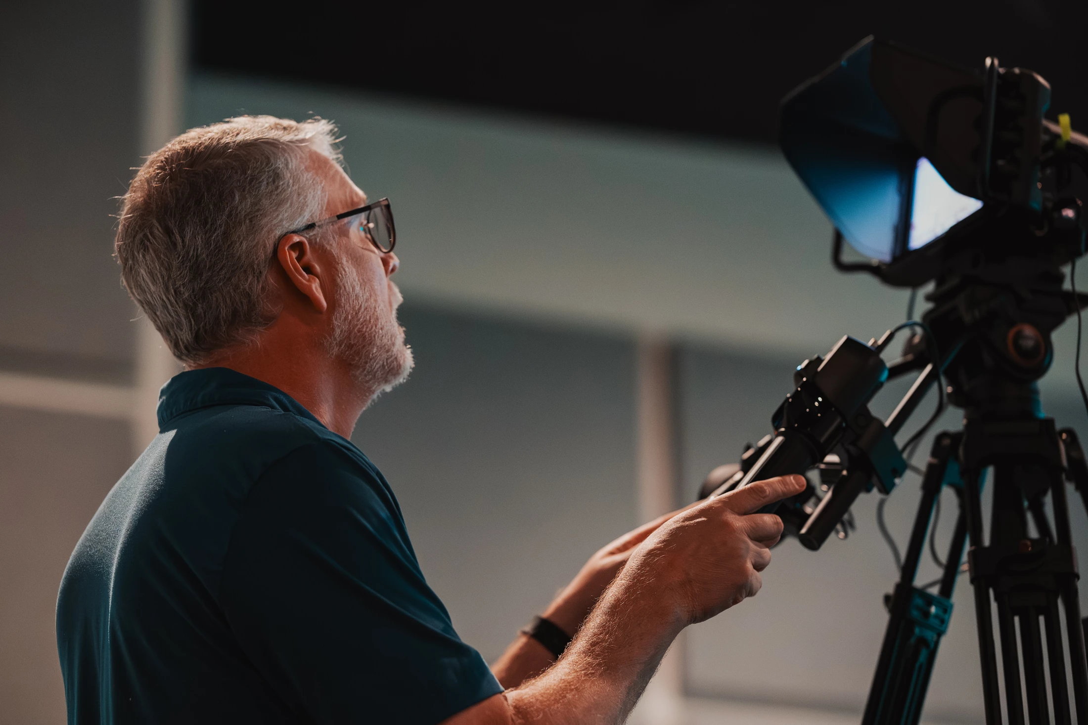 A side profile of a middle-aged man with gray hair and glasses operating a large professional video camera on a tripod in a studio setting.