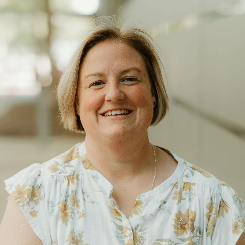 A headshot of a smiling woman with short, blonde hair and freckles. She is wearing a white floral blouse and looking at the camera with a warm expression.
