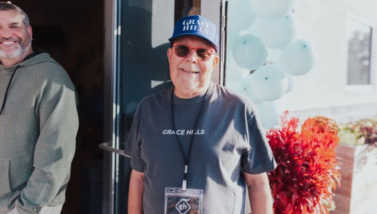 An older man in a Grace Hills hat and shirt smiles at the camera. He's wearing an 'Ask Me' lanyard, standing outside with another man and balloons.