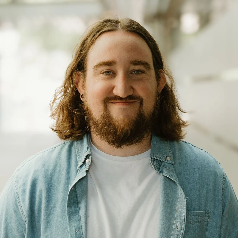 A headshot of a smiling man with long brown hair, a beard, and blue eyes. He is wearing an open blue denim shirt over a white t-shirt.