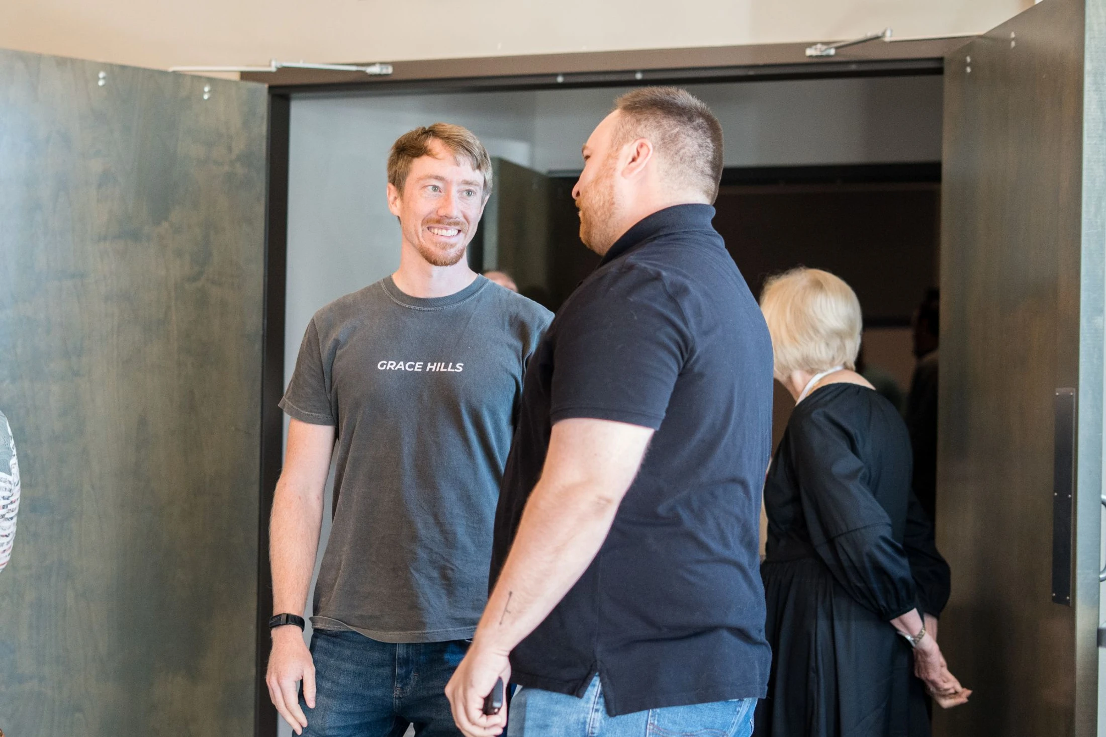A man in a grey 'Grace Hills' t-shirt smiles while talking to another man in a black polo shirt. They are standing indoors near a set of double doors.