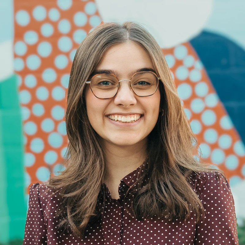 A headshot of a young woman with long brown hair and glasses. She is smiling warmly at the camera, wearing a maroon polka dot shirt against a colorful mural.