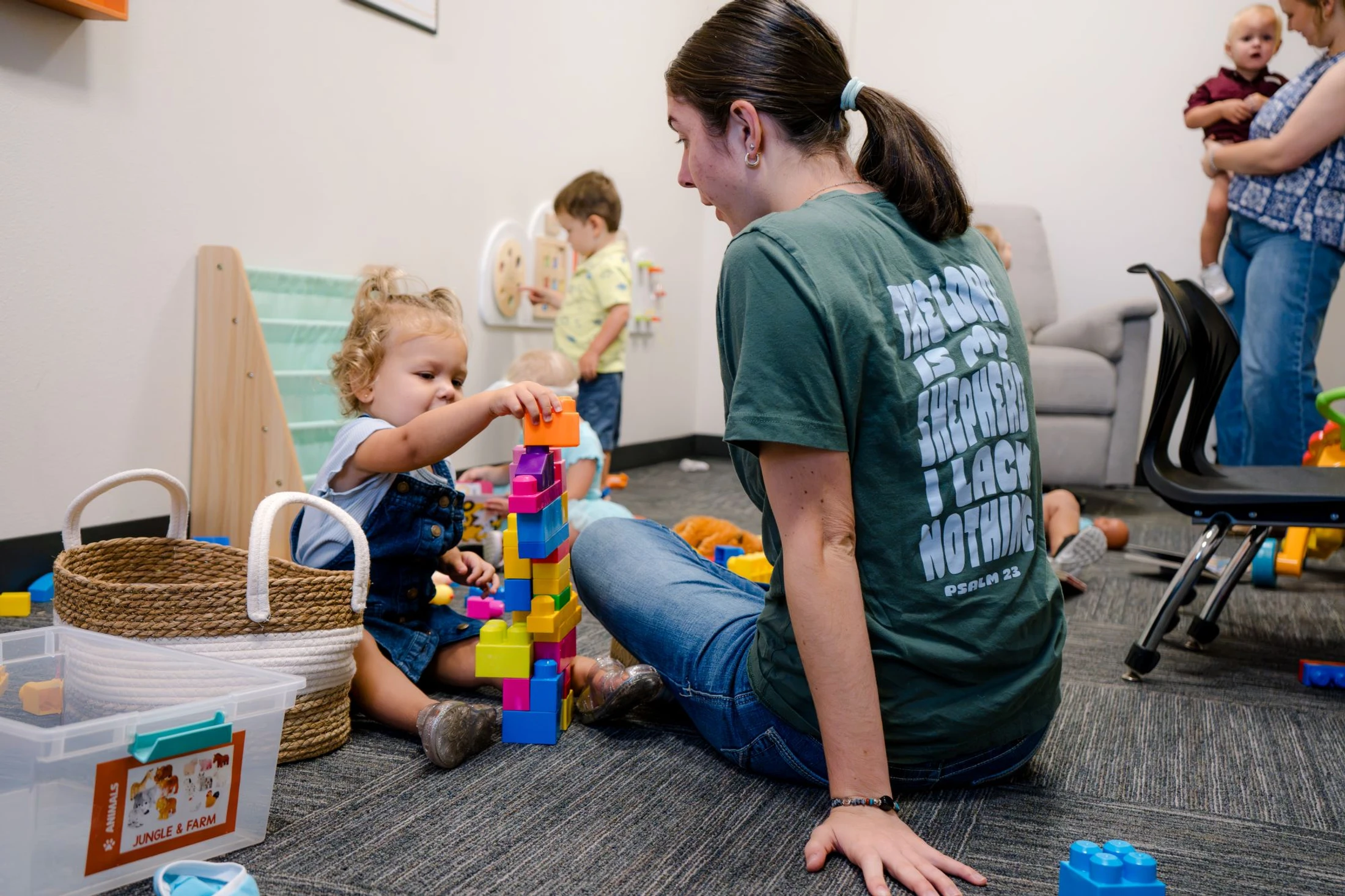 A woman sits on a carpeted floor with a toddler, who is stacking colorful building blocks into a tall tower in a nursery classroom.